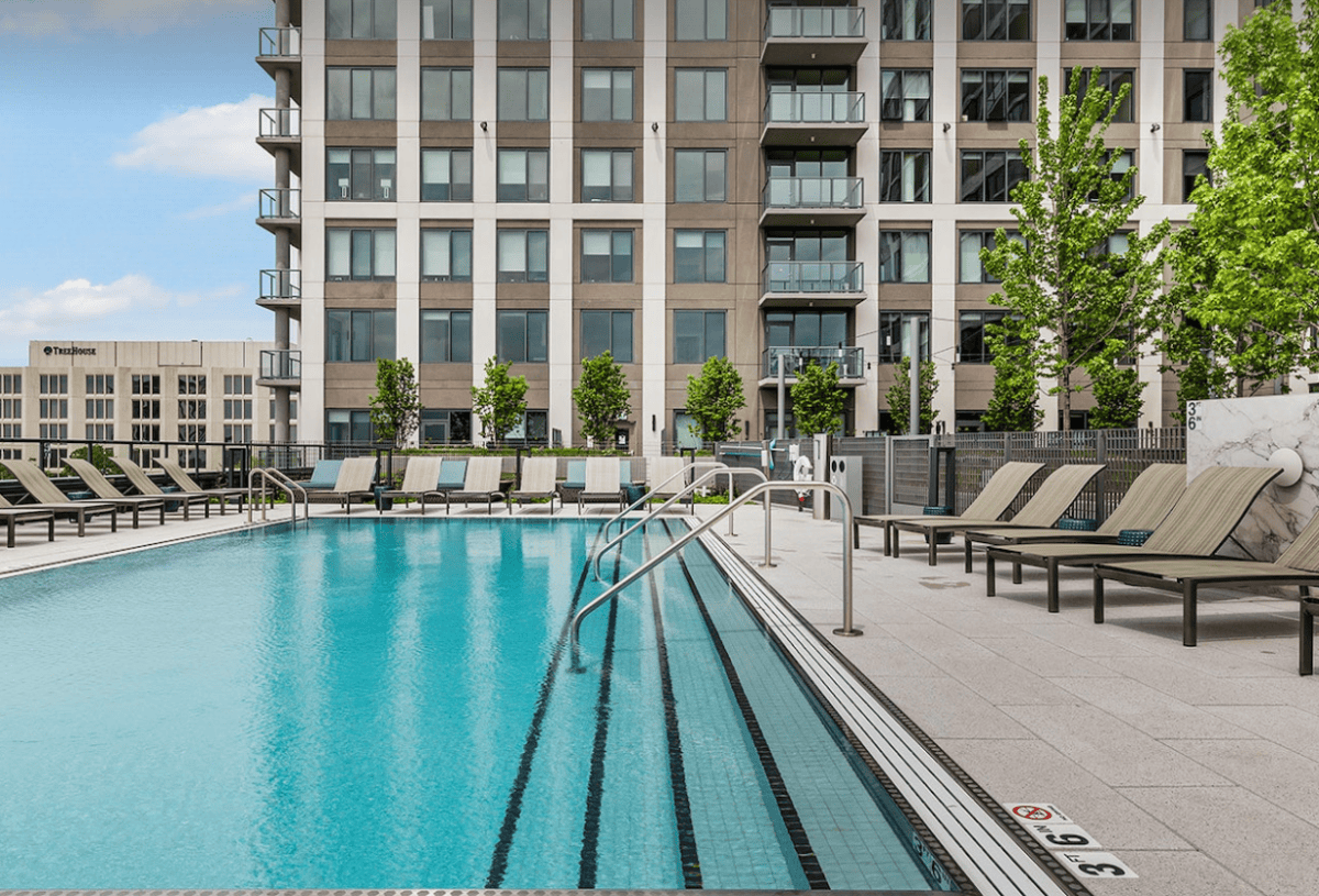 Outdoor swimming pool with metal handrails, surrounded by lounge chairs and trees, adjacent to a modern multi-story apartment building.
