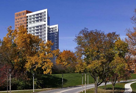 A modern multi-story building rises behind trees with autumn foliage, next to a curved road under a clear blue sky.
