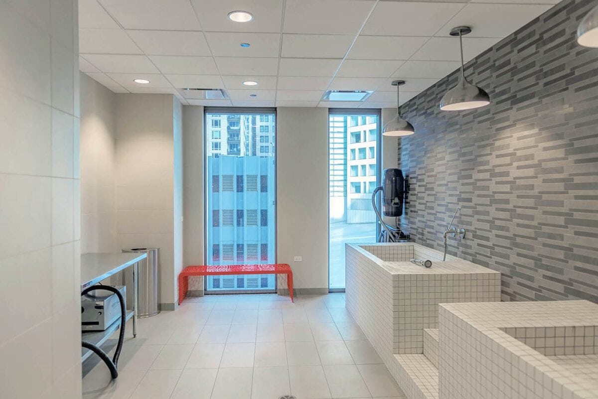 Modern spa room with tiled soaking tubs, gray patterned wall, metal chairs, red bench by floor-to-ceiling windows, and hanging pendant lights.