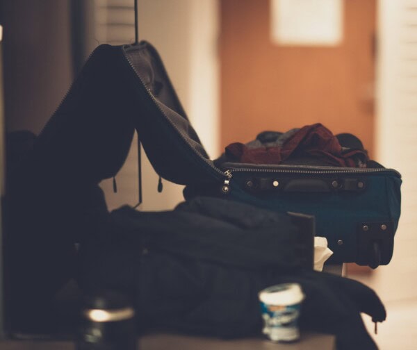 An open suitcase with clothes spilling out sits on a surface next to a yogurt container and a blurred background with a door.
