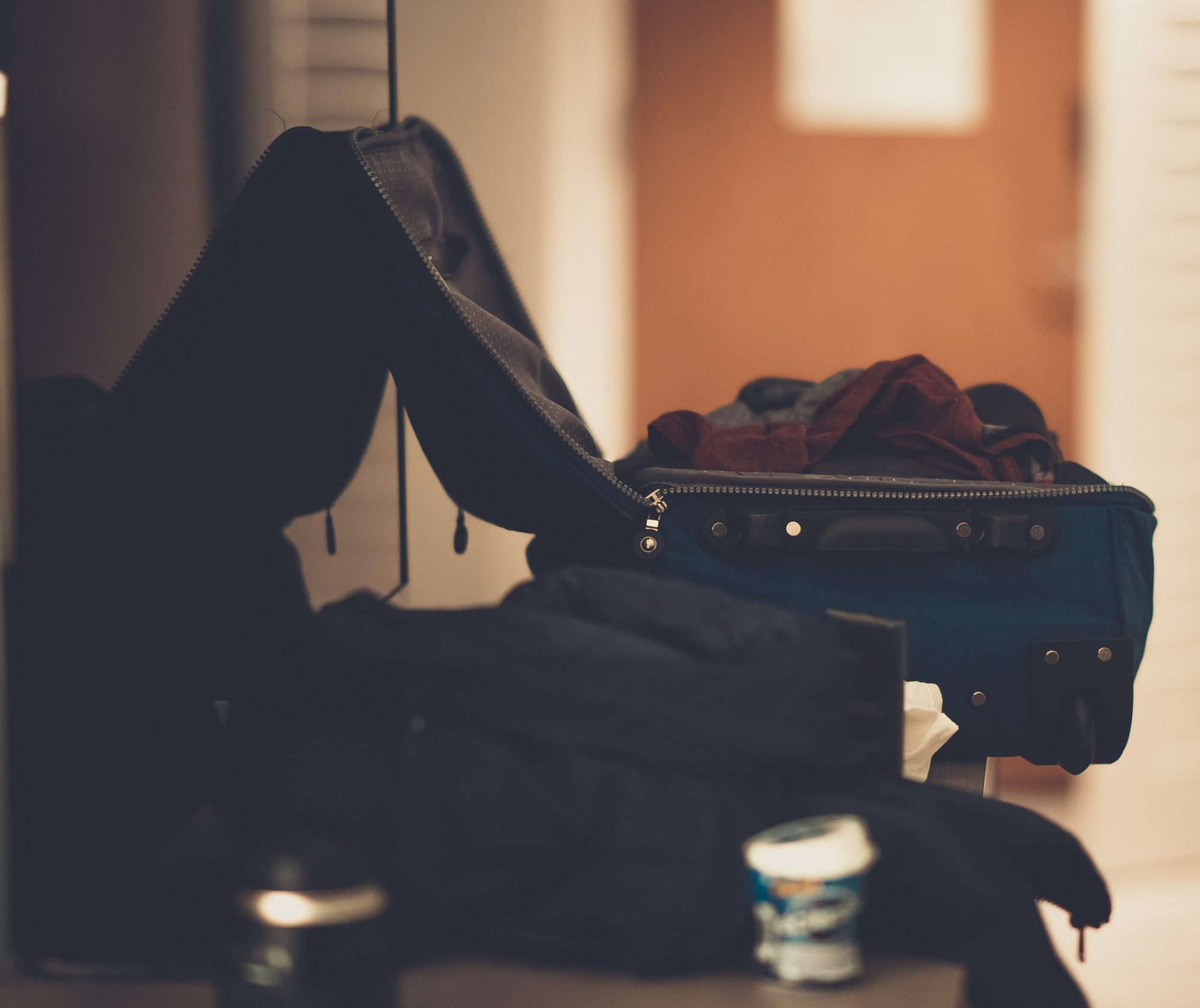 An open suitcase with clothes spilling out sits on a surface next to a yogurt container and a blurred background with a door.