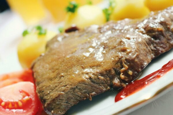 A cooked steak served on a plate with boiled potatoes, sliced tomato, and a side of red sauce.
