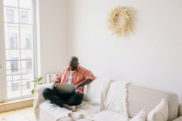 A person sits cross-legged on a light-colored sofa working on a laptop in a bright, minimally decorated room with a large window and a dried wreath on the wall.