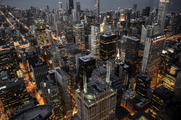 Aerial view of a city skyline at dusk, with tall buildings illuminated and streets grid-patterned with glowing lights.