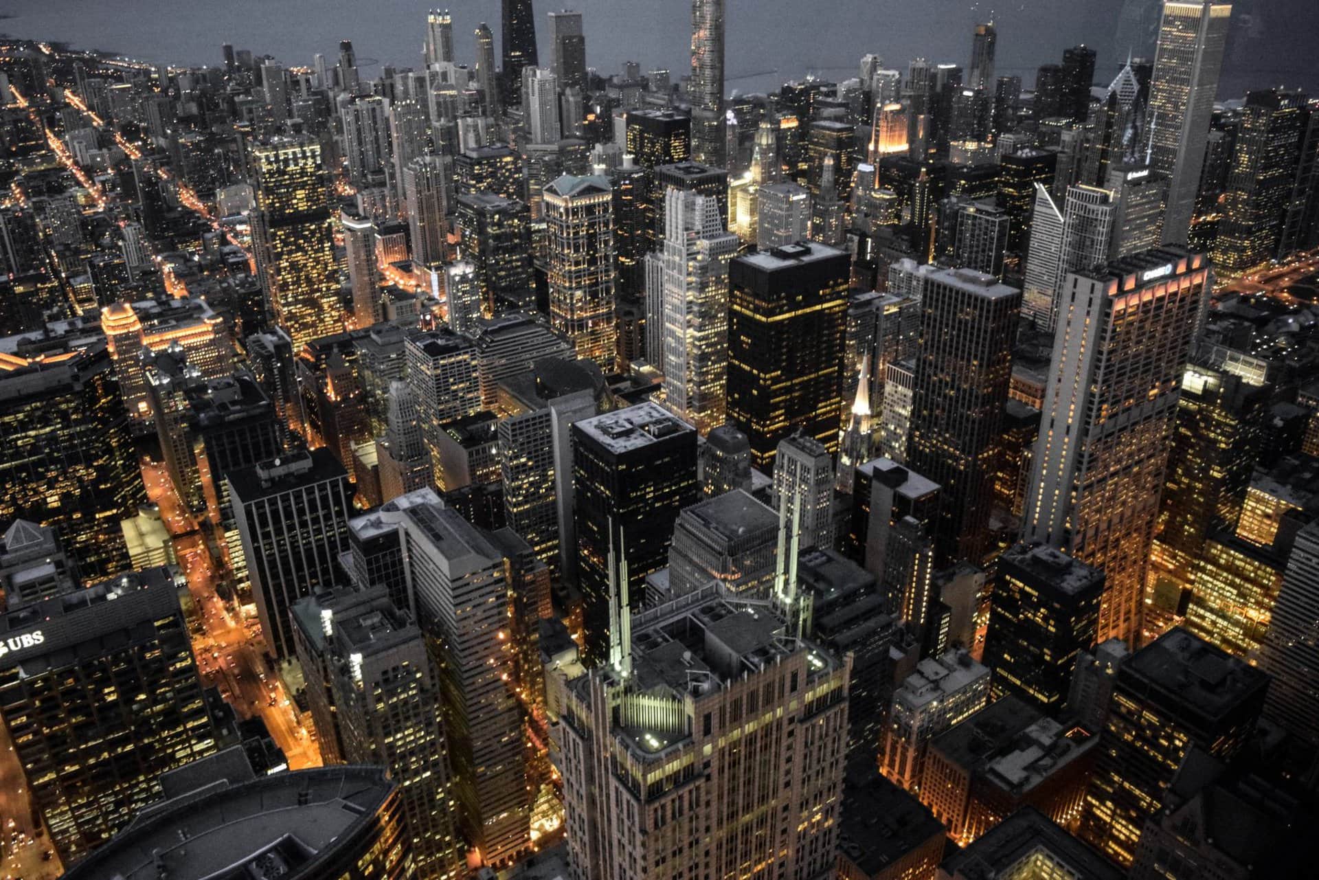 Aerial view of a city skyline at dusk, with tall buildings illuminated and streets grid-patterned with glowing lights.