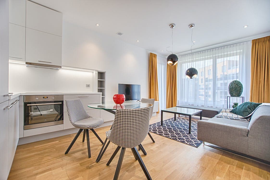 Modern open-plan living room and kitchen with wooden flooring, a gray sofa, glass dining table, patterned chairs, large windows with yellow curtains, and hanging light fixtures.