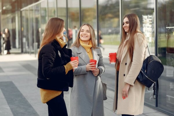 Three women standing outdoors in front of glass windows, holding red cups and talking, dressed in coats and casual clothing.