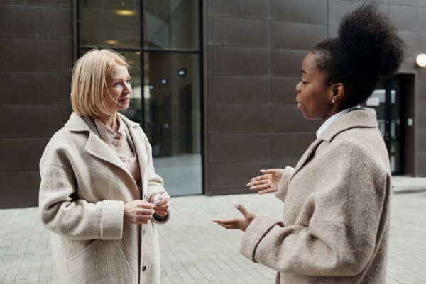 Two women in beige coats stand outside a building, engaged in conversation. One woman gestures with her hands while the other listens attentively.