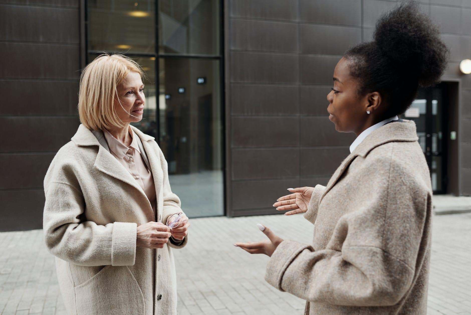 Two women in beige coats stand outside a building, engaged in conversation. One woman gestures with her hands while the other listens attentively.