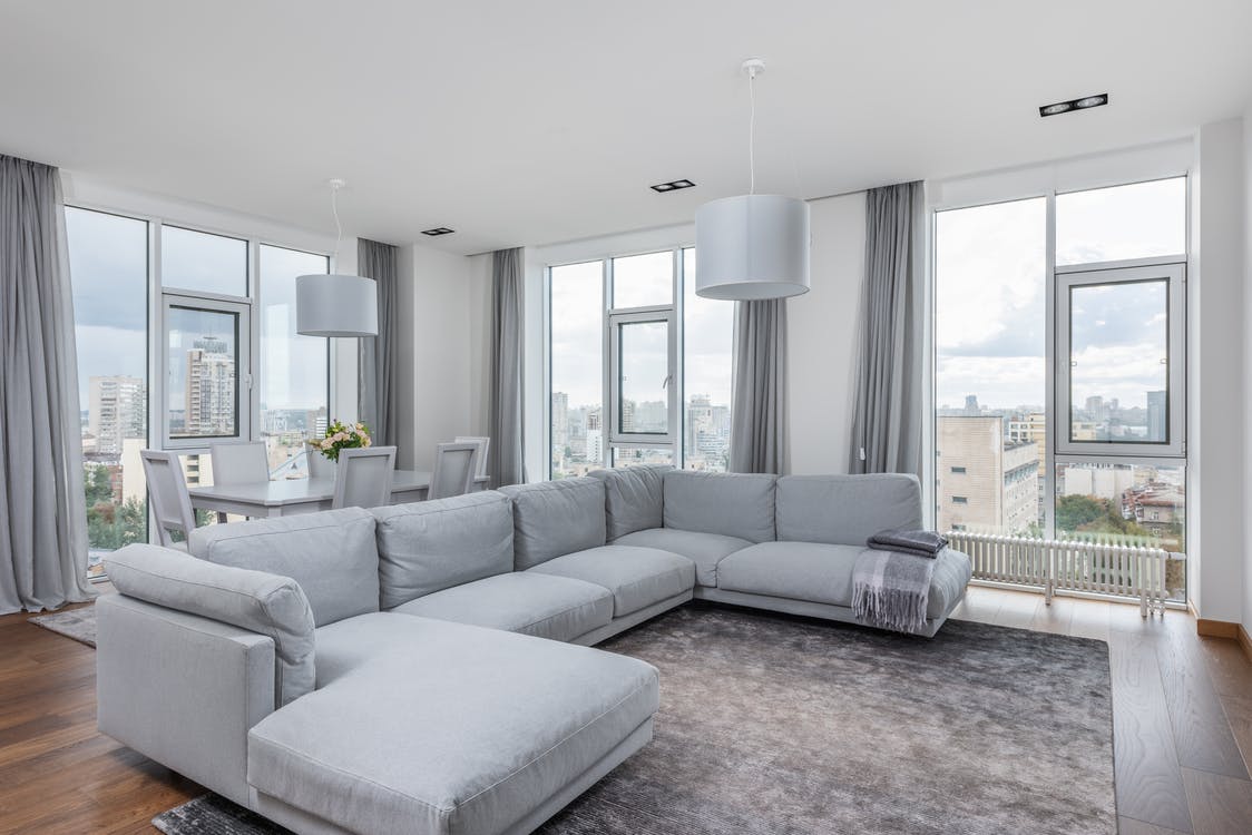 Modern living room with large gray sectional sofa, floor-to-ceiling windows, gray curtains, city views, and a dining area in the background.