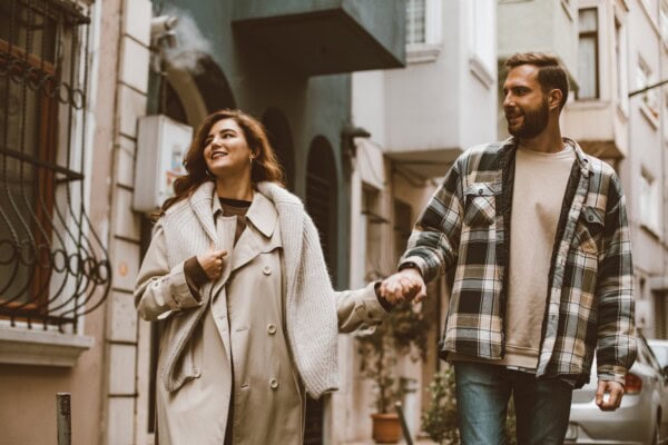 A woman and a man walk hand in hand down a city street, both dressed in casual fall clothing, with buildings and plants in the background.