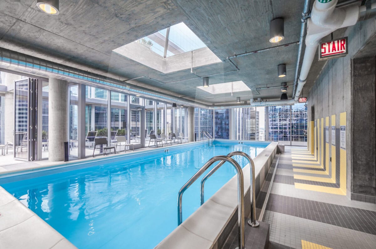 Indoor rooftop pool area with skylights, city views, lounge chairs, concrete ceiling, and a stainless steel ladder leading into the water.