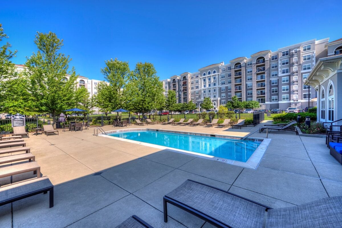 Outdoor swimming pool surrounded by lounge chairs and trees, with multi-story apartment buildings in the background under a clear blue sky.