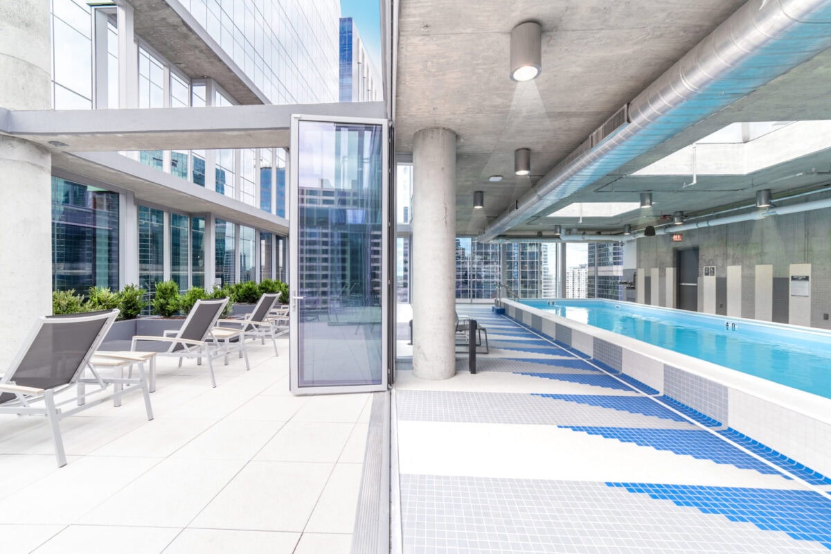 Outdoor rooftop pool with modern lounge chairs, tall concrete columns, and large glass windows overlooking a cityscape.