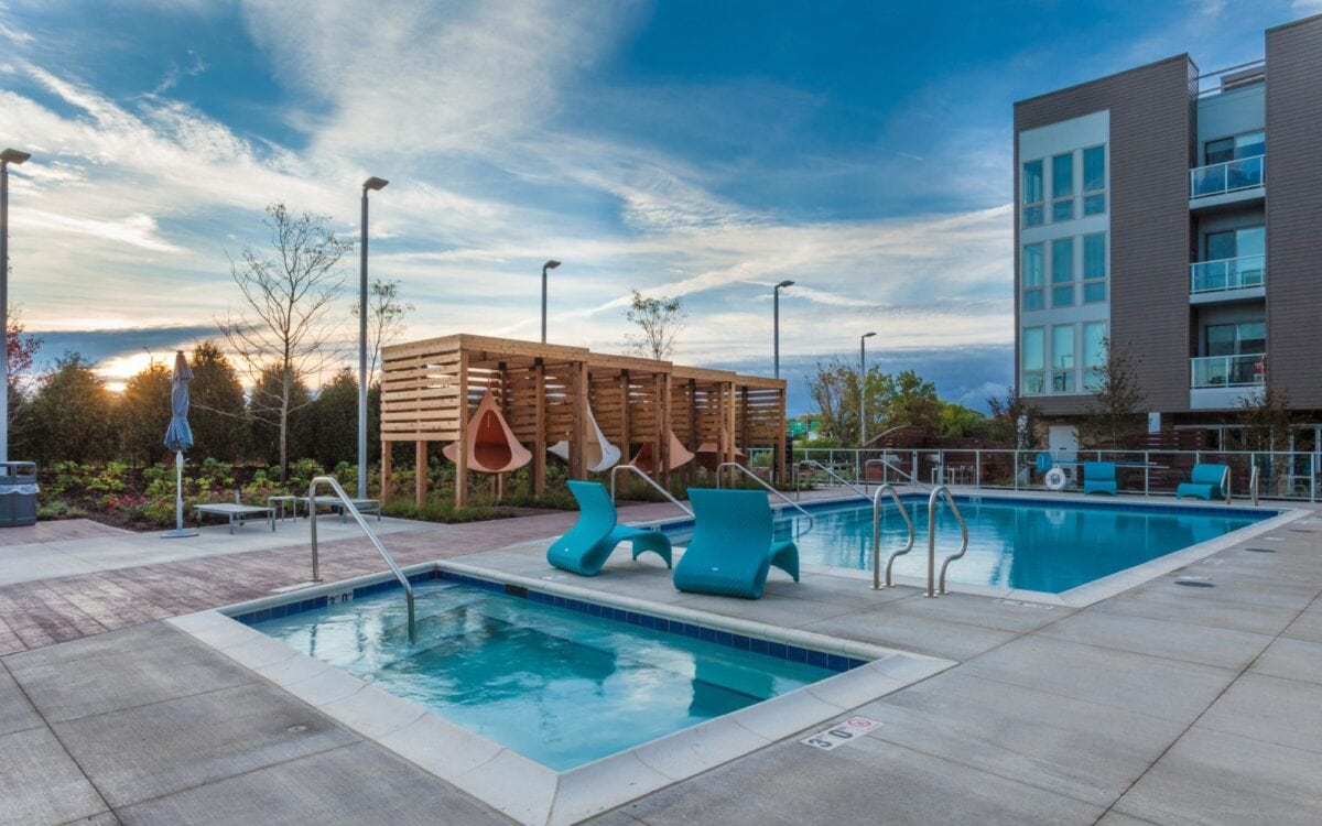 Outdoor pool area with a hot tub, lounge chairs, hanging pod seats, and a modern apartment building in the background under a partly cloudy sky.