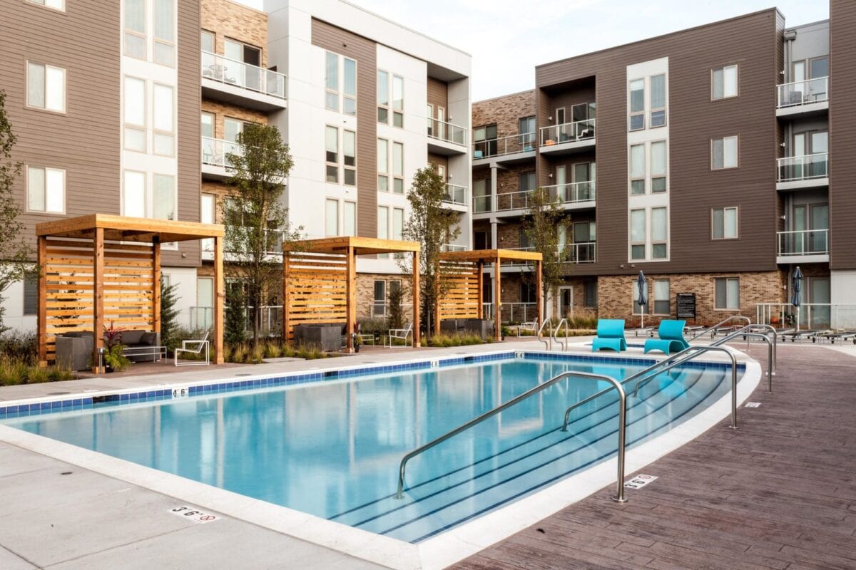 Modern apartment complex with a rectangular outdoor pool, wooden cabanas, poolside chairs, and surrounding multi-story buildings.