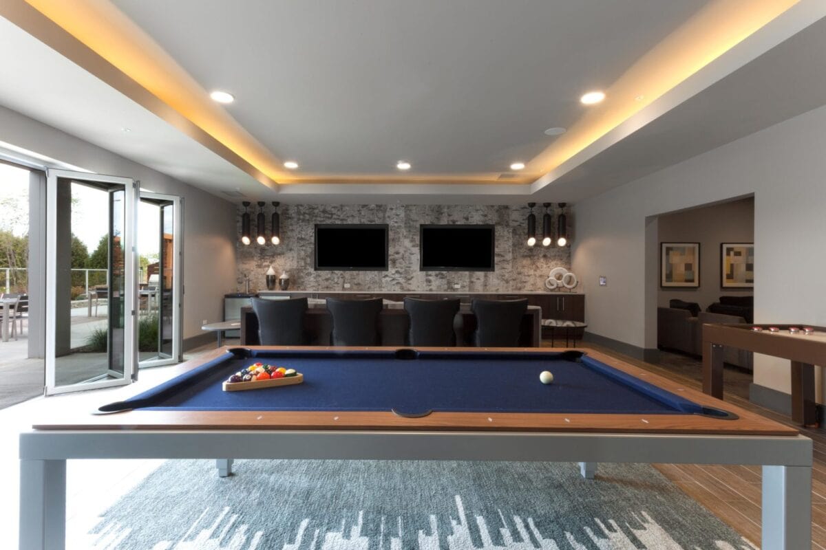 A modern game room with a blue pool table in the foreground, bar seating, two wall-mounted TVs, and large windows opening to the outside.