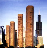 Several tall skyscrapers stand close together against a blue sky, with one dark building featuring antennas among them.