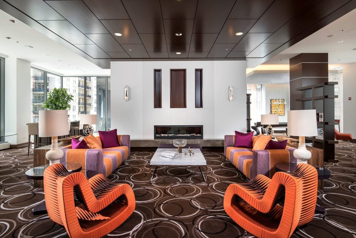 Modern hotel lobby with geometric orange chairs, purple sofas, accent pillows, round patterned carpet, and a fireplace centered on a white wall, surrounded by large windows.
