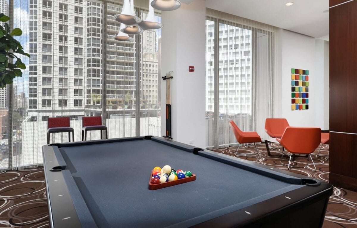 A modern lounge with a black pool table, orange chairs, large windows showing city buildings, and abstract wall art.