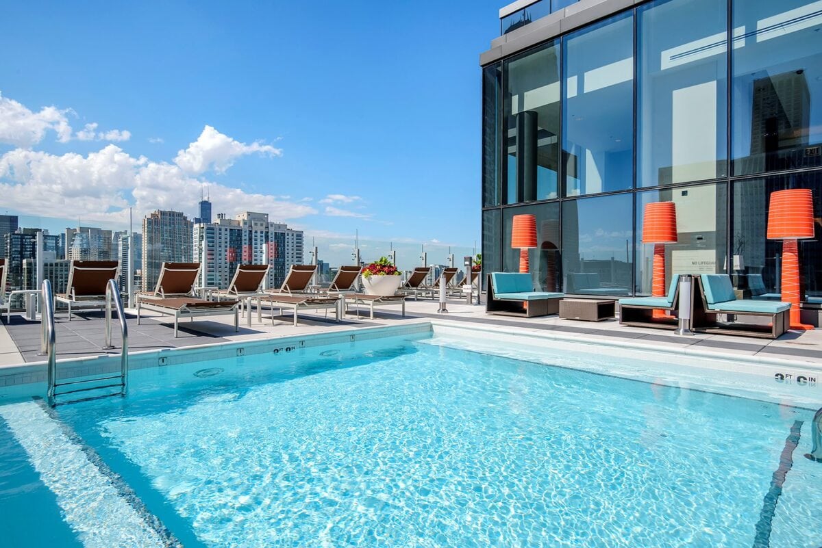 Rooftop outdoor pool with lounge chairs, modern seating, and orange lamps beside a glass building, overlooking a city skyline on a sunny day.