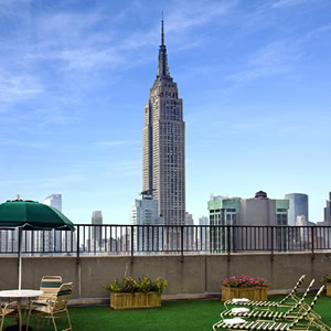 Empire State Building viewed from a rooftop patio with green artificial grass, chairs, tables, potted flowers, and a green umbrella in the foreground.