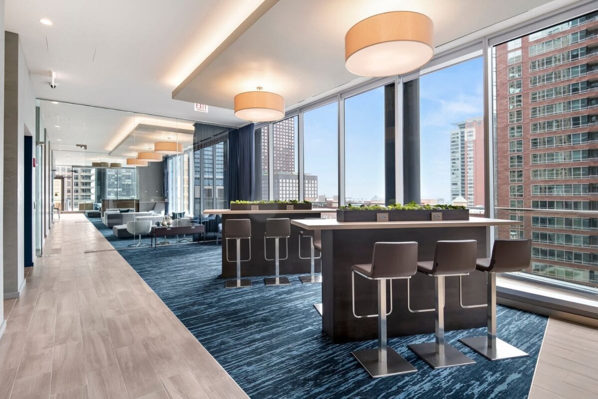 Modern office lounge with tall tables, barstools, large windows, and city views; contemporary lighting and blue carpeting accent the space.