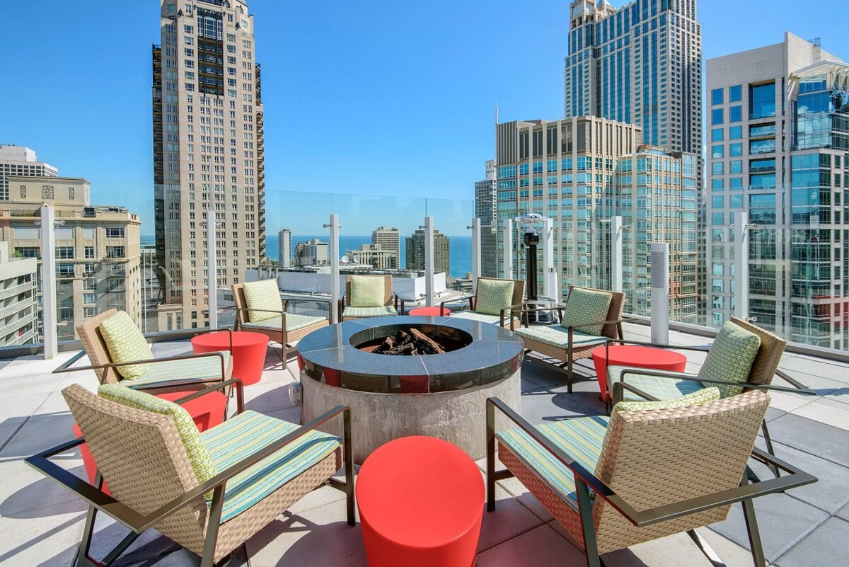 A rooftop patio with a circular fire pit, surrounded by wicker chairs and red stools, set against a city skyline with tall buildings under a clear blue sky.
