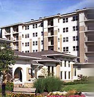 Six-story modern apartment building with white and beige exterior, surrounded by landscaped greenery, flowers, and a bench in the foreground.