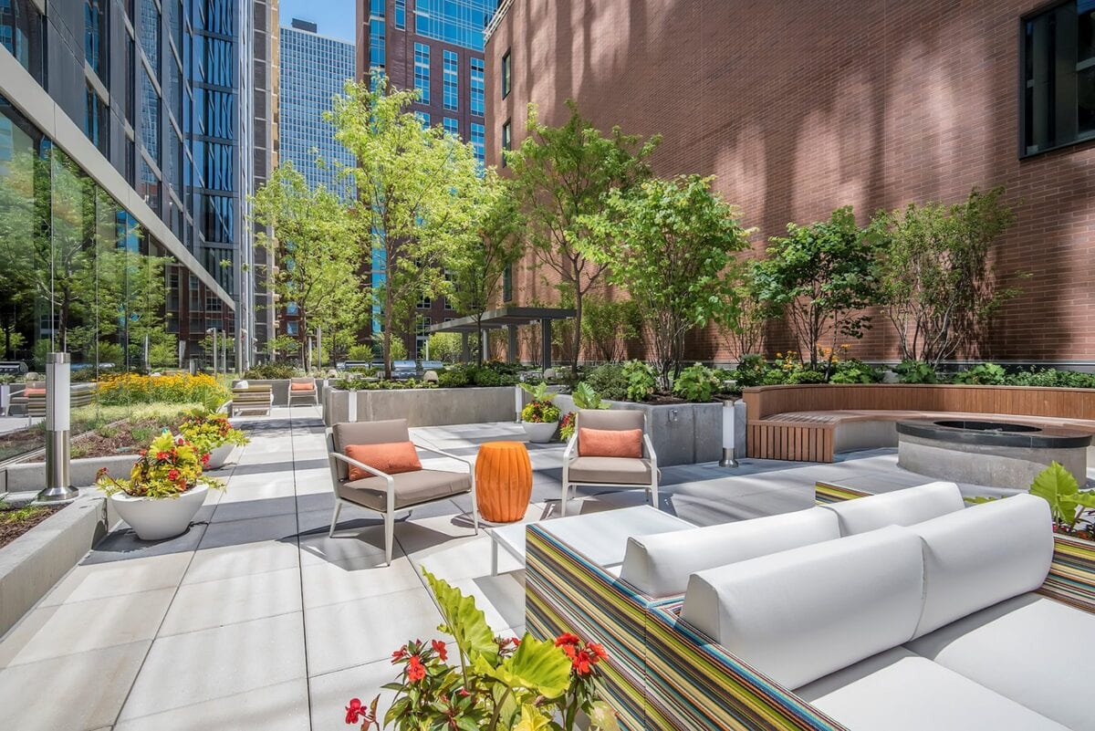 Modern outdoor patio area with white seating, orange and striped cushions, potted plants, trees, and a brick building in the background, surrounded by city skyscrapers.