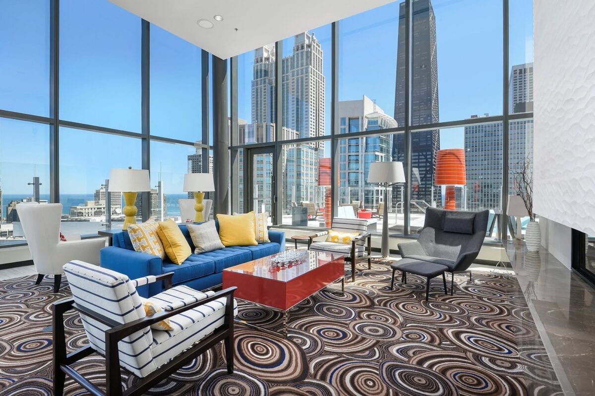 Modern living room with floor-to-ceiling windows, colorful furniture, patterned carpet, and city skyscraper views in the background.