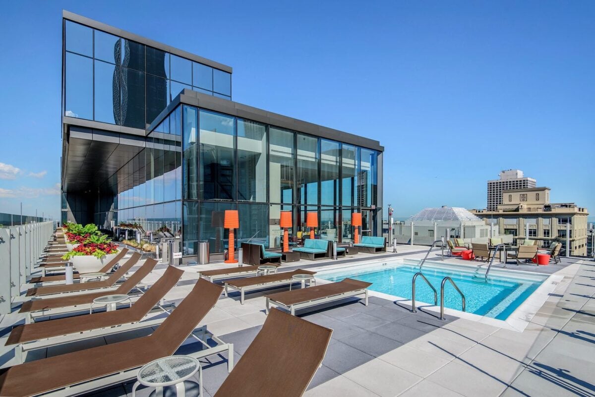 Rooftop pool area with lounge chairs, glass-walled building, potted plants, and cityscape view under a clear blue sky.