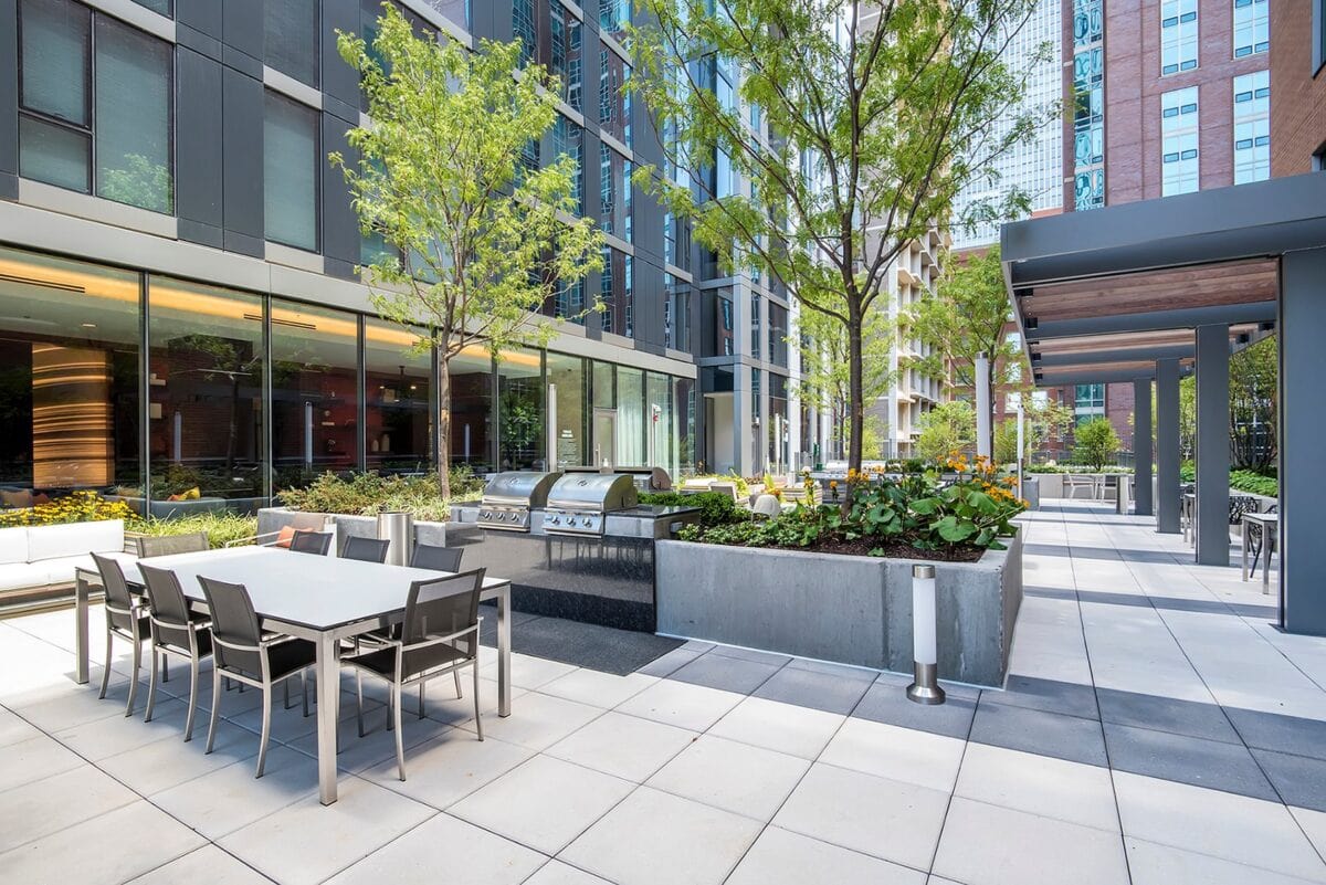 Modern outdoor patio with dining tables, chairs, built-in grills, planters, and trees, surrounded by tall glass buildings.