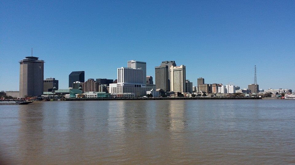 The image shows a city skyline with several tall buildings along a waterfront, seen across a wide river under a clear blue sky.